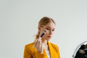 A young woman in a yellow blazer applies makeup with a brush indoors.